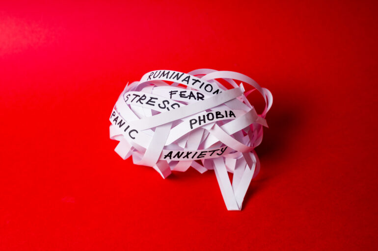 Strips of paper twisted into ball in shape of brain with words STRESS, ANXIETY, PANIC on red background. Tangled thoughts concept, complex mental state