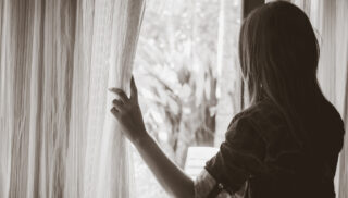 Black and white sad woman holding the curtains open to look out of a large light window at home.