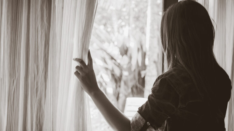 Black and white sad woman holding the curtains open to look out of a large light window at home.