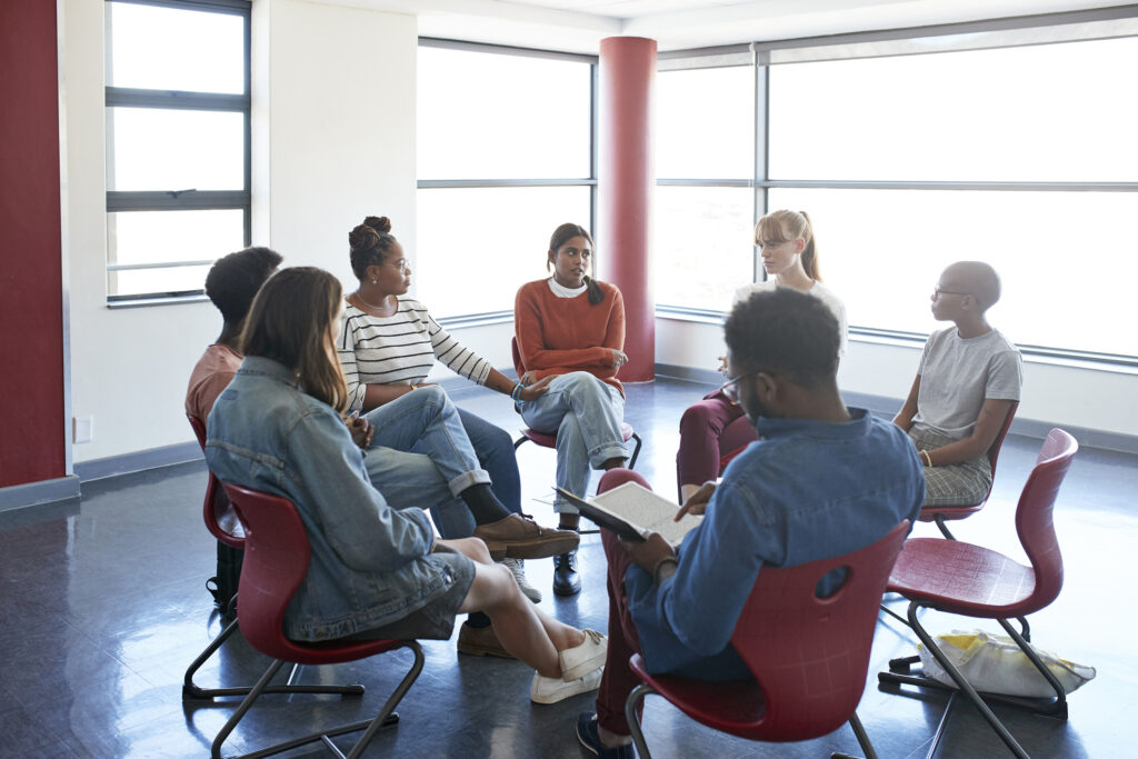 Male mental health professional sitting amidst young multi-ethnic students during psychotherapy session in classroom