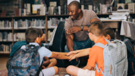 Elementary school teacher huddling with a group of kids in a library. Male educator motivating his students in a primary school. Academic motivation and mentorship.