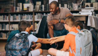 Elementary school teacher huddling with a group of kids in a library. Male educator motivating his students in a primary school. Academic motivation and mentorship.