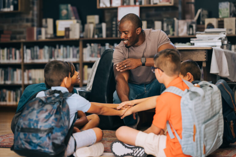 Elementary school teacher huddling with a group of kids in a library. Male educator motivating his students in a primary school. Academic motivation and mentorship.