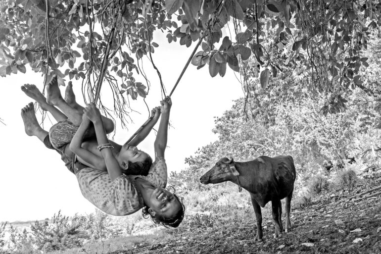 Photo courtesy of Vital Impacts BIHAR, India: Children play on makeshift swings tied to trees on a river island, finding moments of joy in a landscape reshaped each year by seasonal floods. Photographer Afzal Khan’s long-term project, The Other Side, documents daily life on these low-lying islands, among the most flood-prone regions in the world, where families continually adapt to a shifting river and an uncertain future.