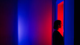 A woman stands in profile, bathed in vibrant red light from an open doorway while surrounded by deep blue shadows. The themes of decision-making and the courage to step into the unknown.
