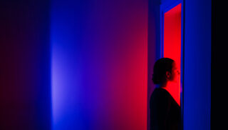A woman stands in profile, bathed in vibrant red light from an open doorway while surrounded by deep blue shadows. The themes of decision-making and the courage to step into the unknown.