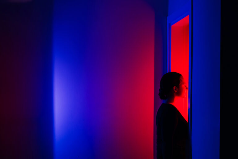 A woman stands in profile, bathed in vibrant red light from an open doorway while surrounded by deep blue shadows. The themes of decision-making and the courage to step into the unknown.
