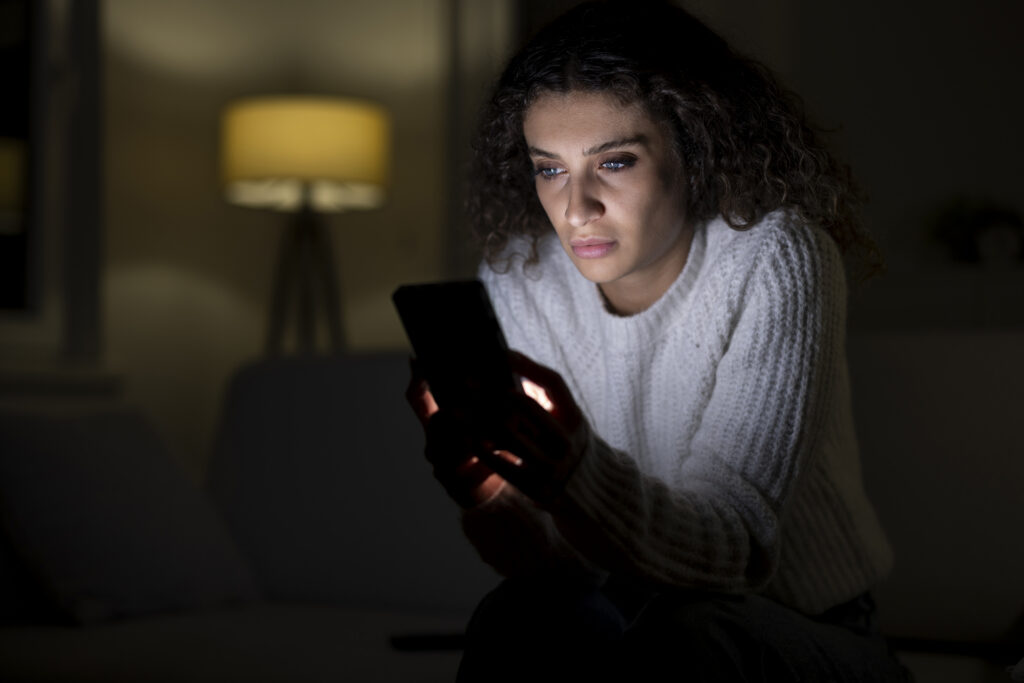 A dramatic portrait of a serious young woman sitting in a dark living room late at night. Her face is intensely illuminated by the glow of her smartphone screen as she reads messages, highlighting themes of insomnia, bad news, or digital addiction.